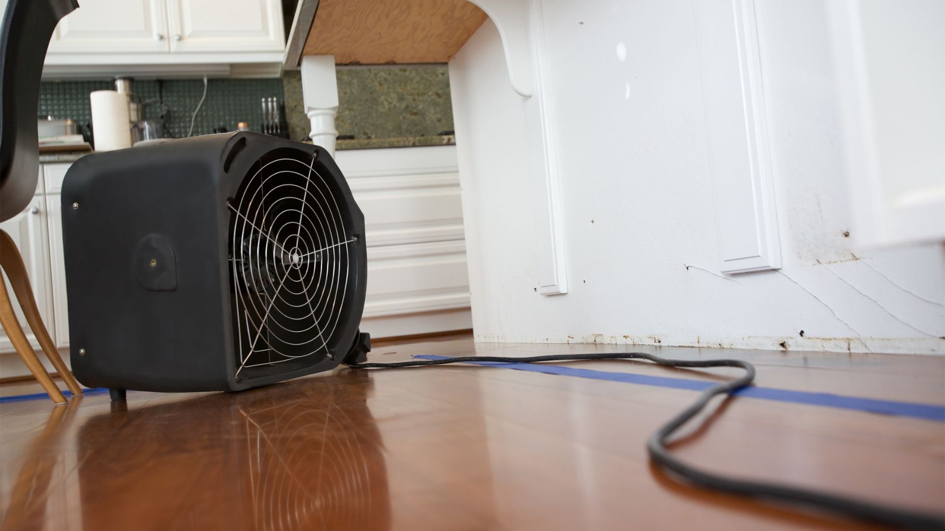 Black industrial fan on wooden floor near kitchen with power cord